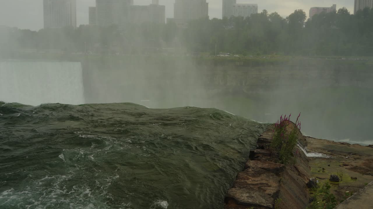 Close-up view of water rushing over the edge of Niagara Falls with mist and distant city skyline in the background