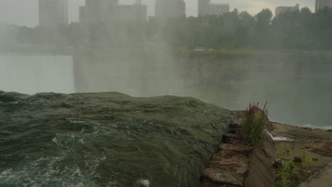 close-up view of water rushing over the edge of niagara falls with mist and distant city skyline in the background