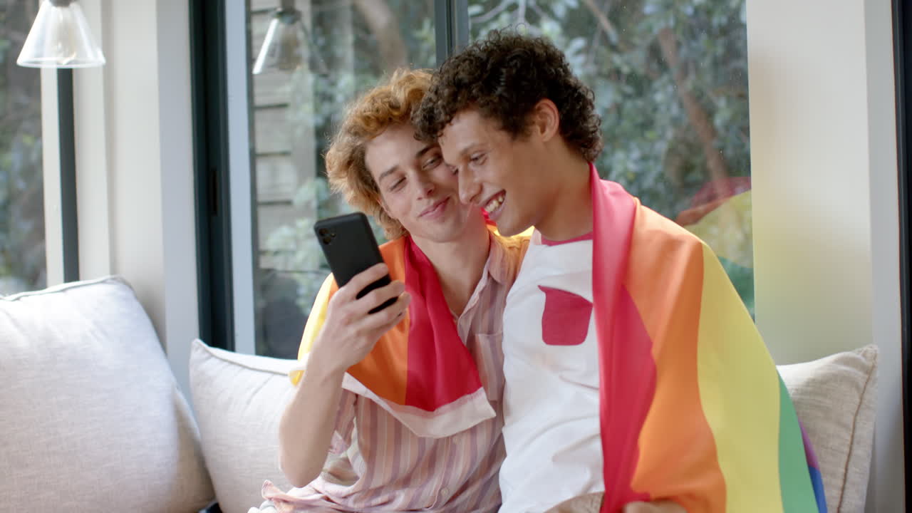 Diverse gay male couple with rainbow lgbt flag, using smartphone on sofa at home, slow motion