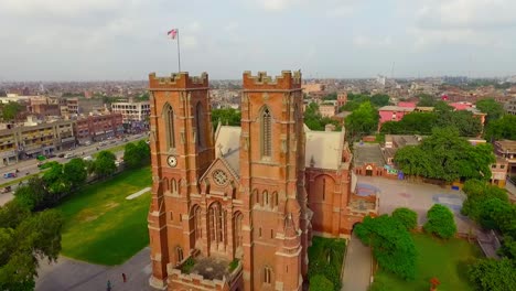 Luftaufnahme-Einer-Schönen-Alten-Kirche-Mit-Stadt-Im-Hintergrund,-Schöne-Bäume-Und-Gras-Rund-Um-Die-Kirche,-Tauben,-Die-über-Die-Kirche-Fliegen,-Menschen,-Die-Sich-Außerhalb-Der-Kirche-Bewegen