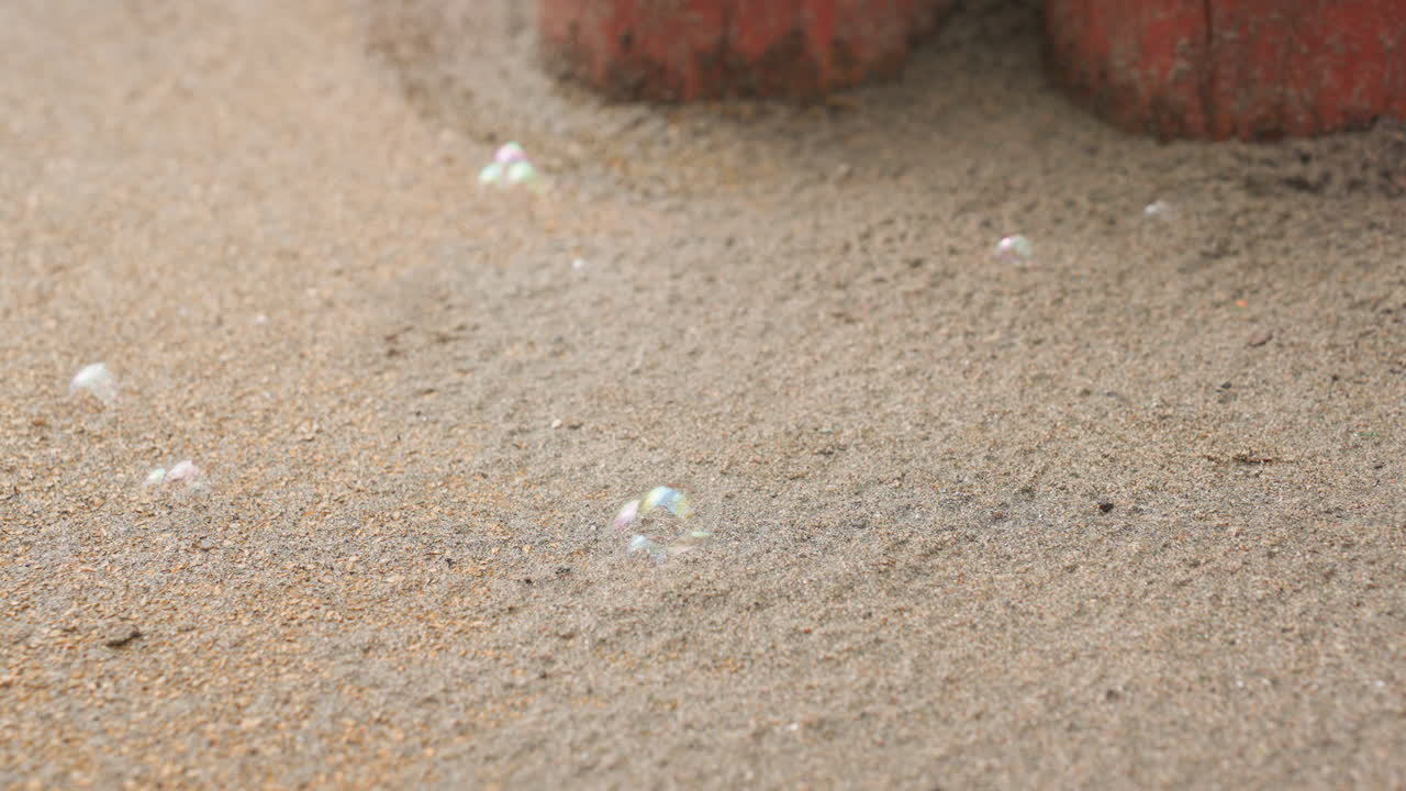 Close up of single soap bubble on sandy ground as wind pushes surface, wobbling and sliding across grains near worn red post, subtle shimmer with rainbow reflections under soft