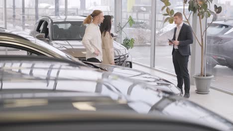 young car salesman showing to young couple new automobile at dealership salon.