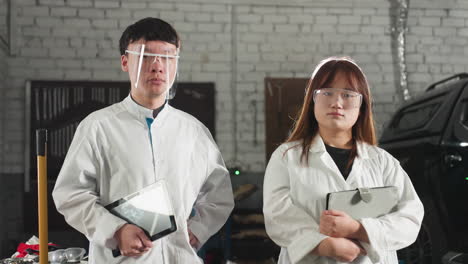 two students in lab coats stand near an engine in mechanical workshop, holding tablets while focused on their work, protective eyewear and industrial setting