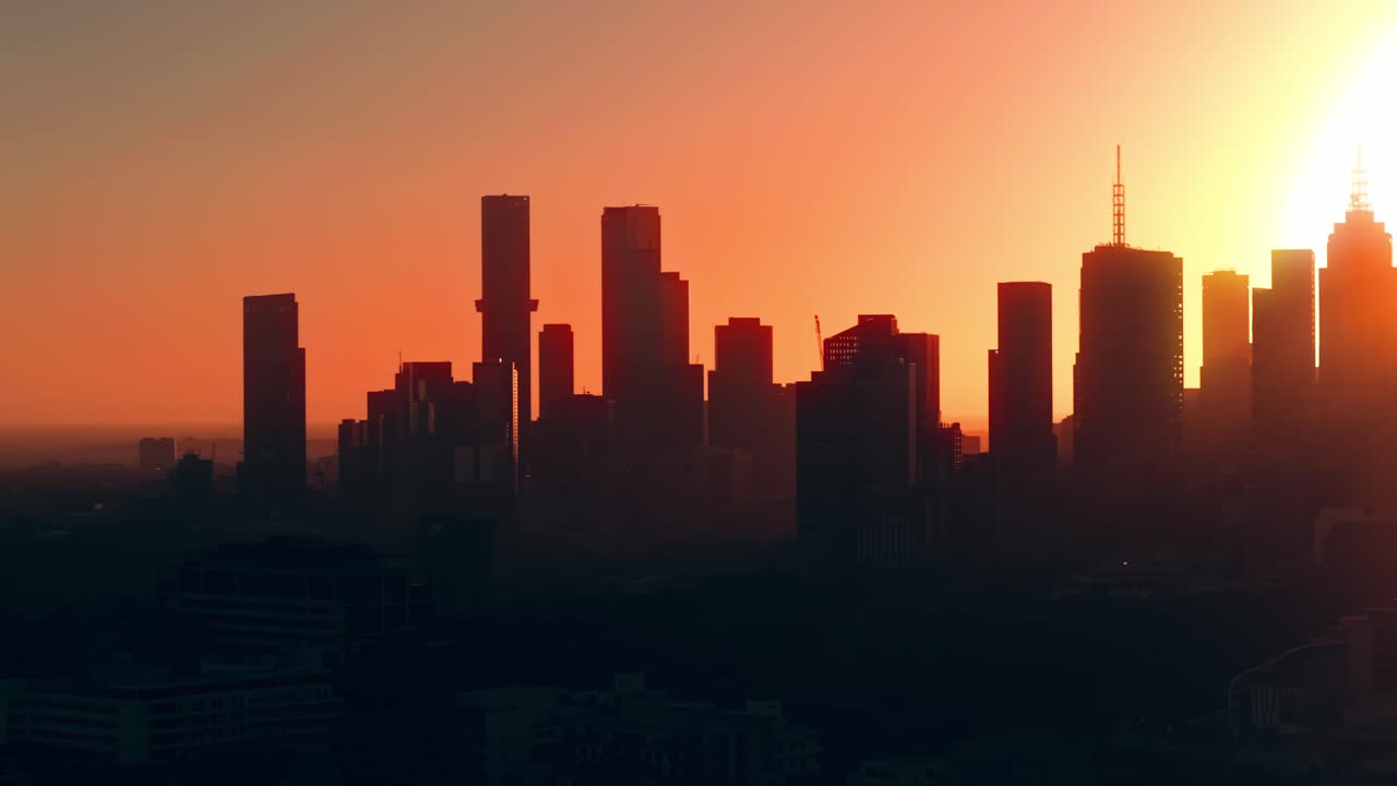 Cálido resplandor vespertino sobre el horizonte de la ciudad de Melbourne, seguimiento aéreo hacia la derecha, silueta abstracta de edificios con textura natural.