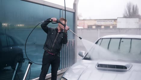 Young-man-in-leather-jacket-washing-his-silver-sport-car-at-self-service-car-wash.-High-pressure-water-coming-out.-Slowmotion-shot