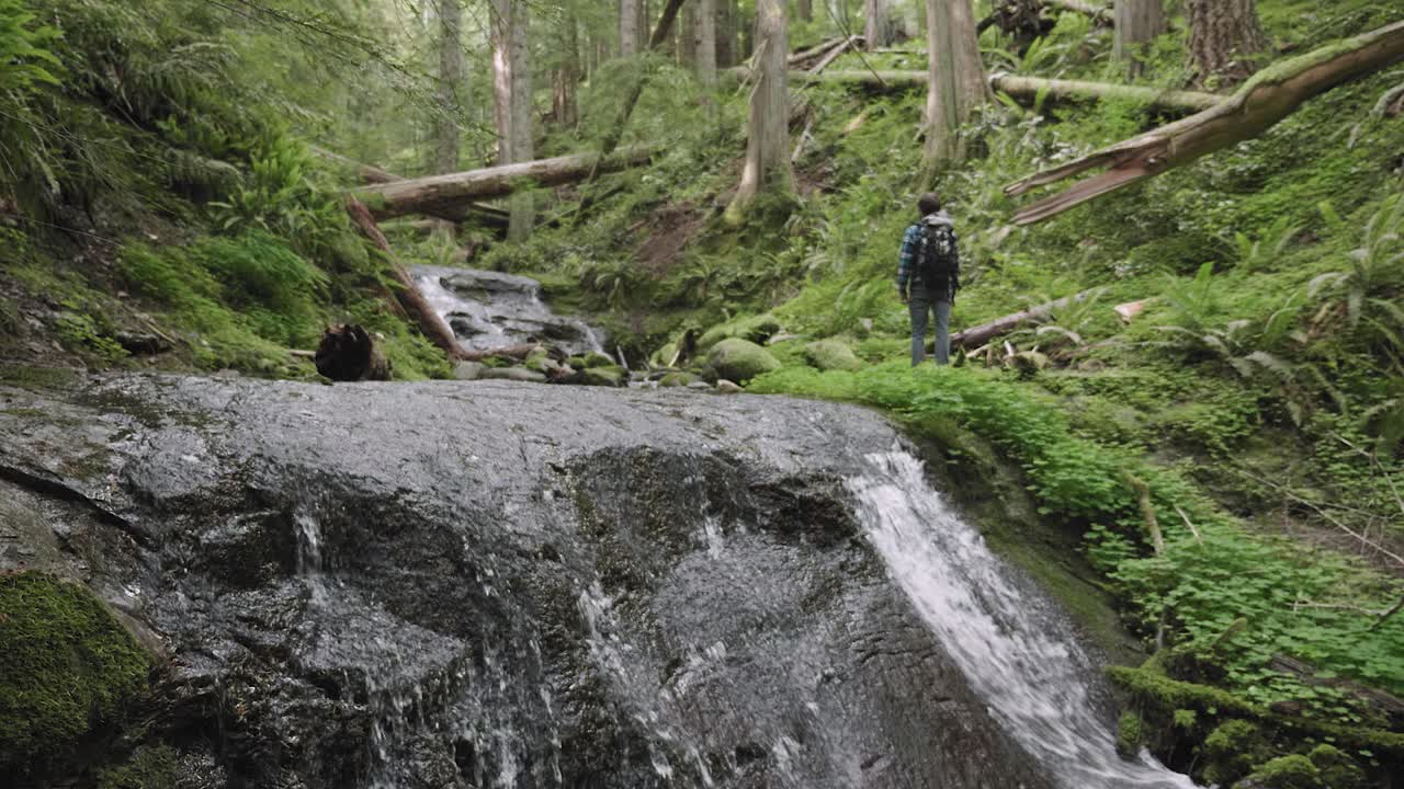 White male hiking through peaceful lush green forest in the distance with double waterfalls rushing stopping to enjoy the atmosphere