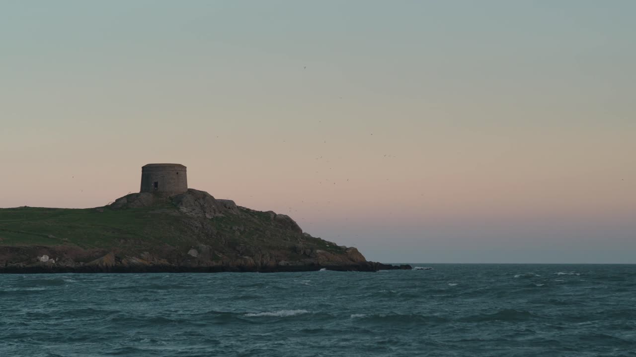 A locked-off shot of Dalkey Island’s Martello Tower in Dublin, Ireland. A flock of birds soars behind the historic stone tower as waves ripple under a pastel sunset sky.