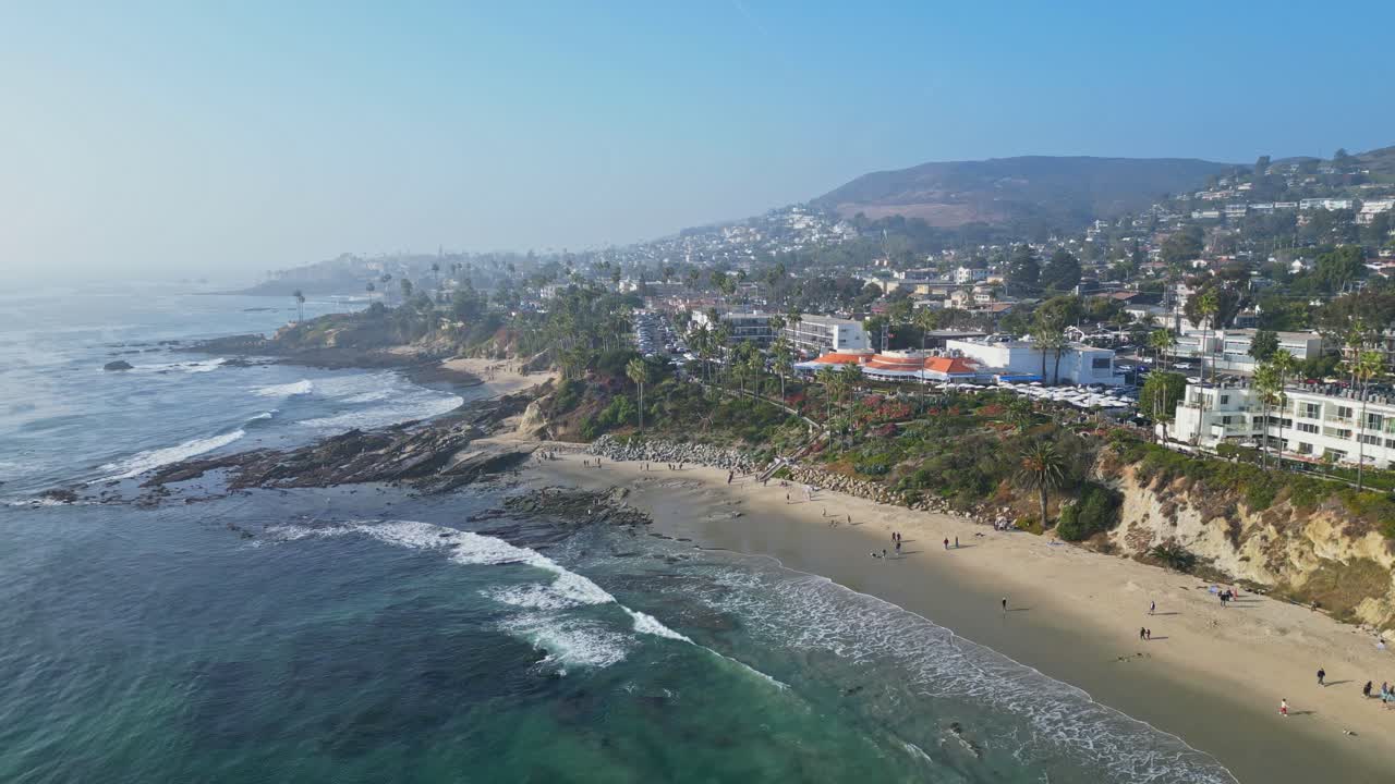 A drone shot zooming in on Laguna Beach, capturing crashing waves, sunlight, and people enjoying the shore.
