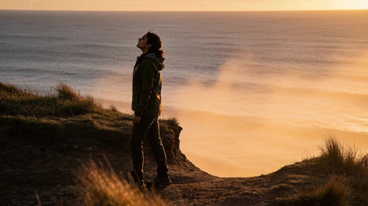 Walking hiker on cliff-path slowing watching warm horizon mist in green hooded jacket boots