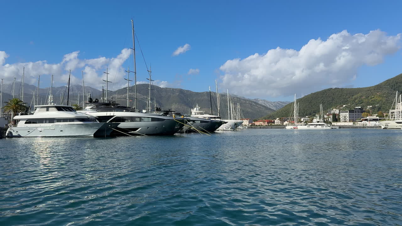 Vista panoramica di yacht di lusso e barche a vela ancorati a Porto Montenegro, incorniciati da acque calme e blu, montagne e un cielo terso e limpido. Atmosfera da porticciolo Premium, ideale per viaggi, lifestyle e turismo.