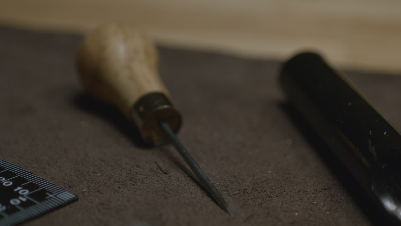 Detailed view of a sharp leatherworking awl with a polished wooden handle, lying on a brown surface beside a ruler and black tool.