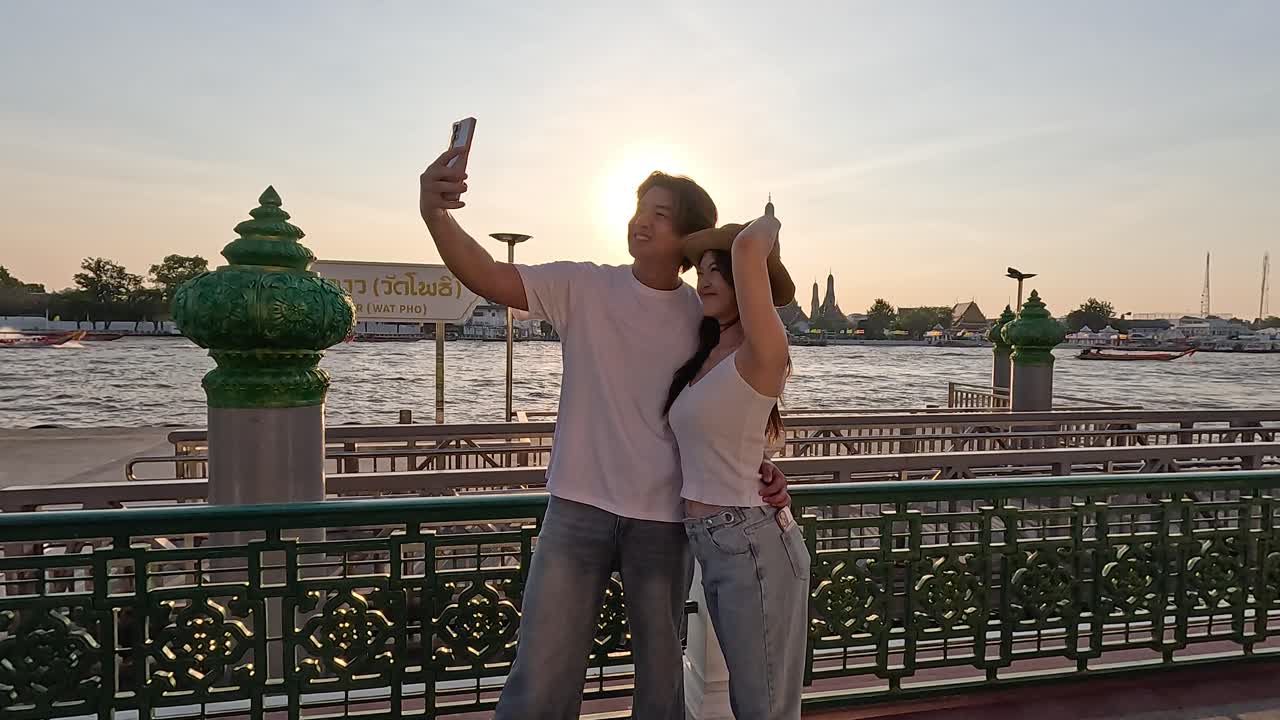 Una coppia sorridente scatta un selfie con lo smartphone sul lungofiume durante il tramonto nell'ora d'oro, movimento della fotocamera a mano libera
