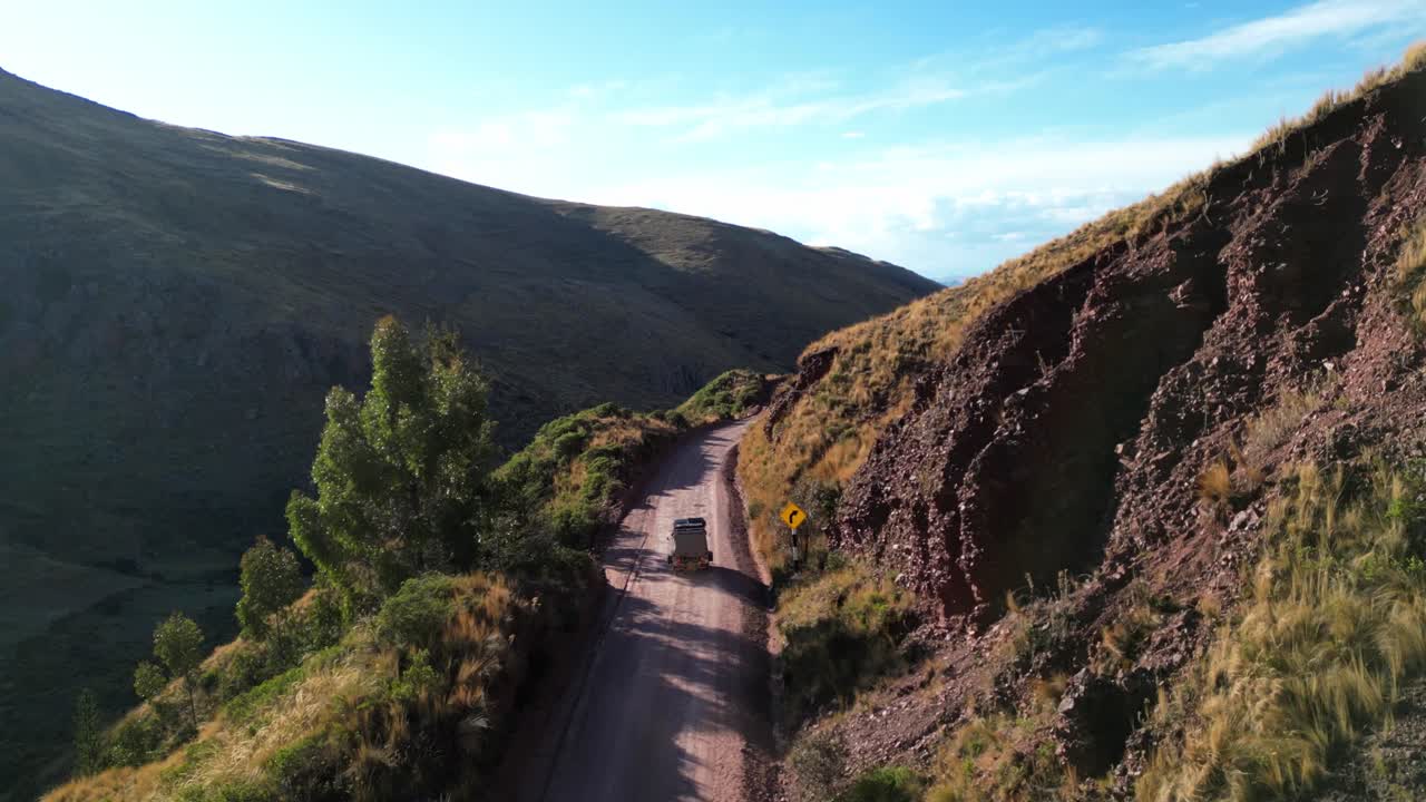 Cinematic drone follow shot of a tuk-tuk travelling a mountain road in the Andes. The golden light of sunset glows across rugged peaks and valleys