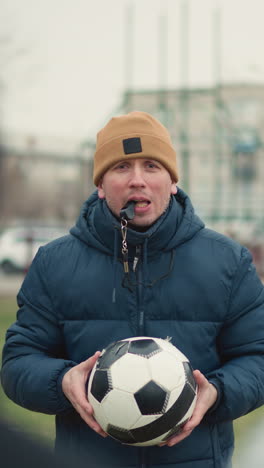 close-up of a coach instructing two boys with a whistle in his mouth, holding a soccer ball, the coach throws the ball at the person wearing a gray top