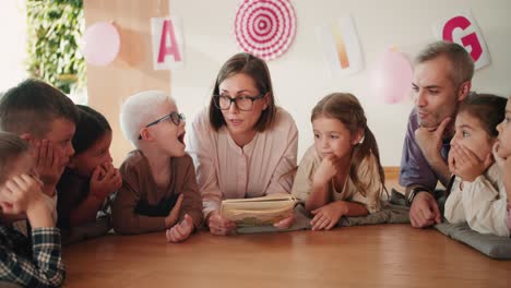 Eine-Lehrerin-Mit-Bob-Frisur,-Brille-Und-Weißem-Hemd-Liegt-Mit-Den-Kindern-Auf-Speziellen-Kissen-Auf-Dem-Boden-Und-Liest-Ihnen-In-Ihrer-Ersten-Unterrichtsstunde-Zur-Vorbereitung-Auf-Die-Schule-Im-Schulvorbereitungsclub-Der-Kinder-Ein-Buch-Vor