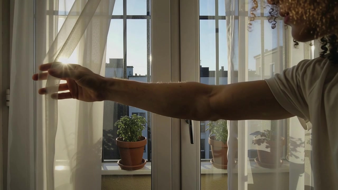 Sun urging man short-sleeve light shirt pulling sheer drape at balcony door showing pots