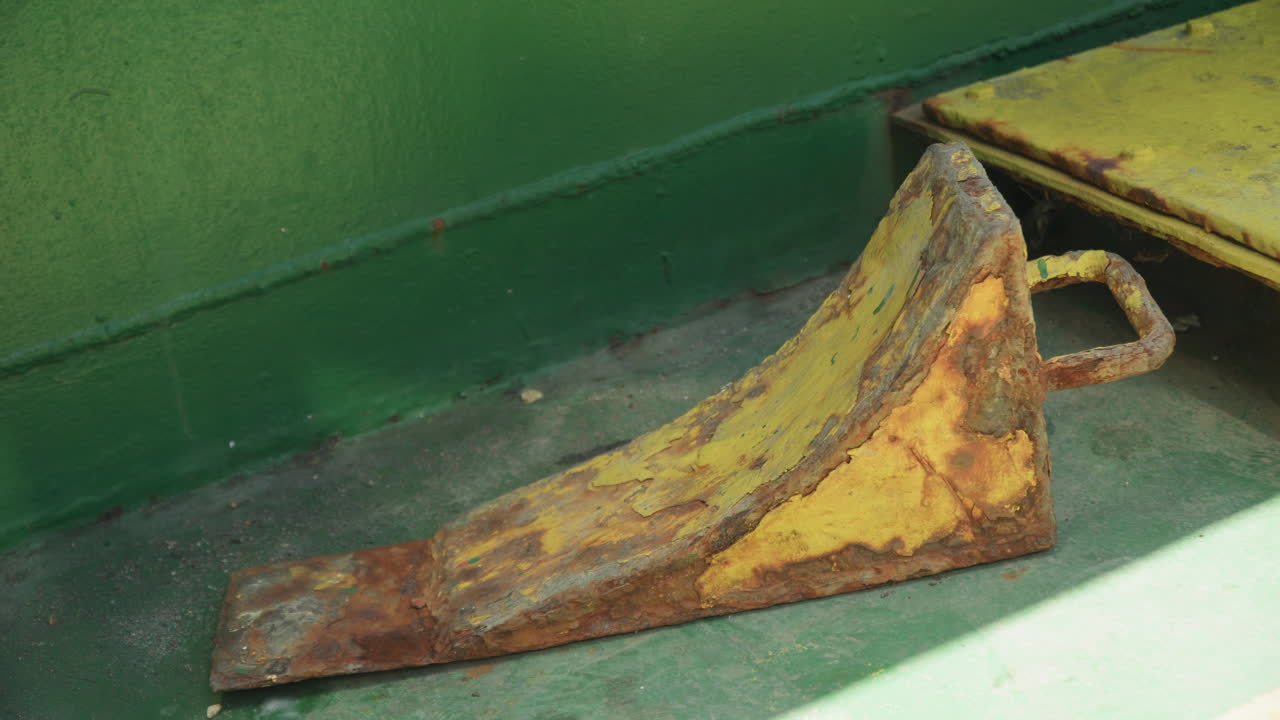 Rusten gul hjulkile på dæk med afskallende maling og kraftig oxidation, nærbillede af korroderet kile og løkkehåndtag mod falmet grøn skot formidler forladt skibsværftsatmosfære med bjærgningsarbejder
