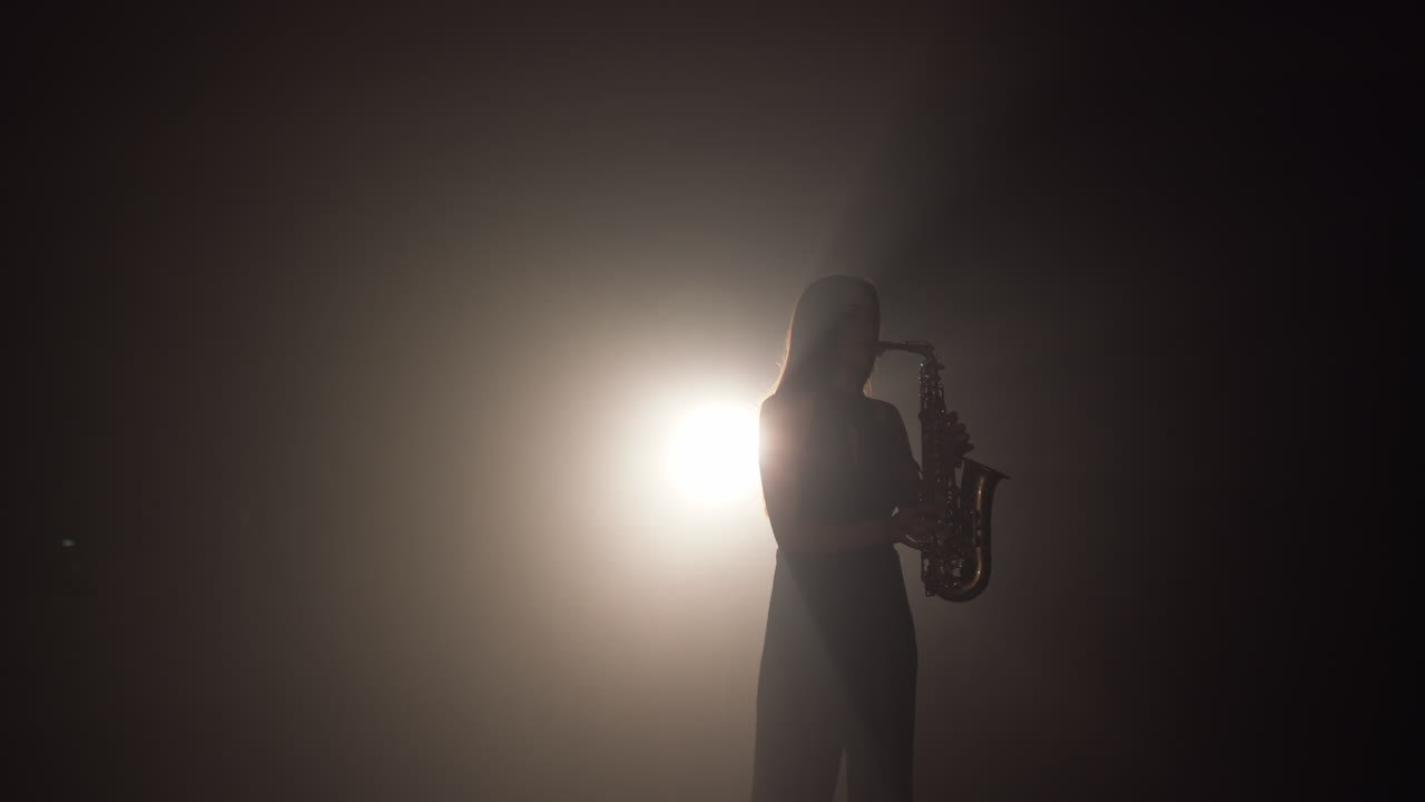 Young beautiful girl in a dark dress plays on a golden shiny saxophone on stage dark studio with