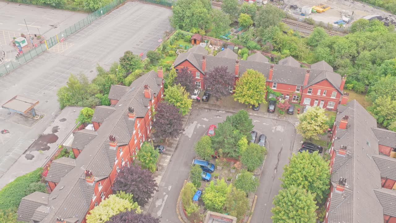 Drone push-in over red brick terraced houses in Salford, showing an inner courtyard with trees, parked cars, and gardens near railway tracks and industrial lots
