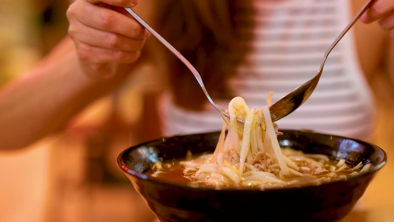 A person skillfully twirls noodles with a fork and spoon in a warmly lit Gold Coast restaurant, capturing a moment of culinary enjoyment