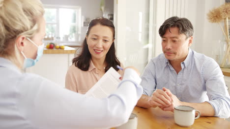 성숙한 아시아 커플이 집에서 팬데 기간 동안 마스크를 입은 여성 금융 자문가와 만나고 있습니다.