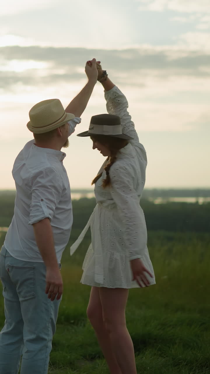 한 남자가 평온한 호수 의 잔디 언덕에서 아내를 회전시니다. 색 셔츠, 모자, 청바지를 입은 남자는 색 드레스와 검은색 모자를 입은 여자가 그를 따뜻하게 포옹합니다.