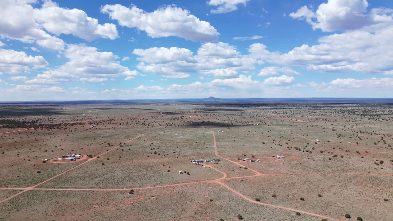 Drone flying forward over the desert of Williams Arizona, south of the Grand Canyon. Clouds are dotting the sky over warm, dusty dirt on a bright sunny day. There are dirt roads and a few structures.