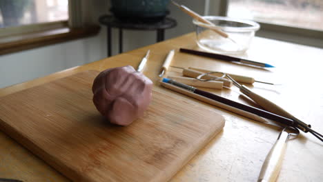 a ball of brown modeling clay for sculpting, tools, brushes and art supplies on a wooden table in an art studio slide left