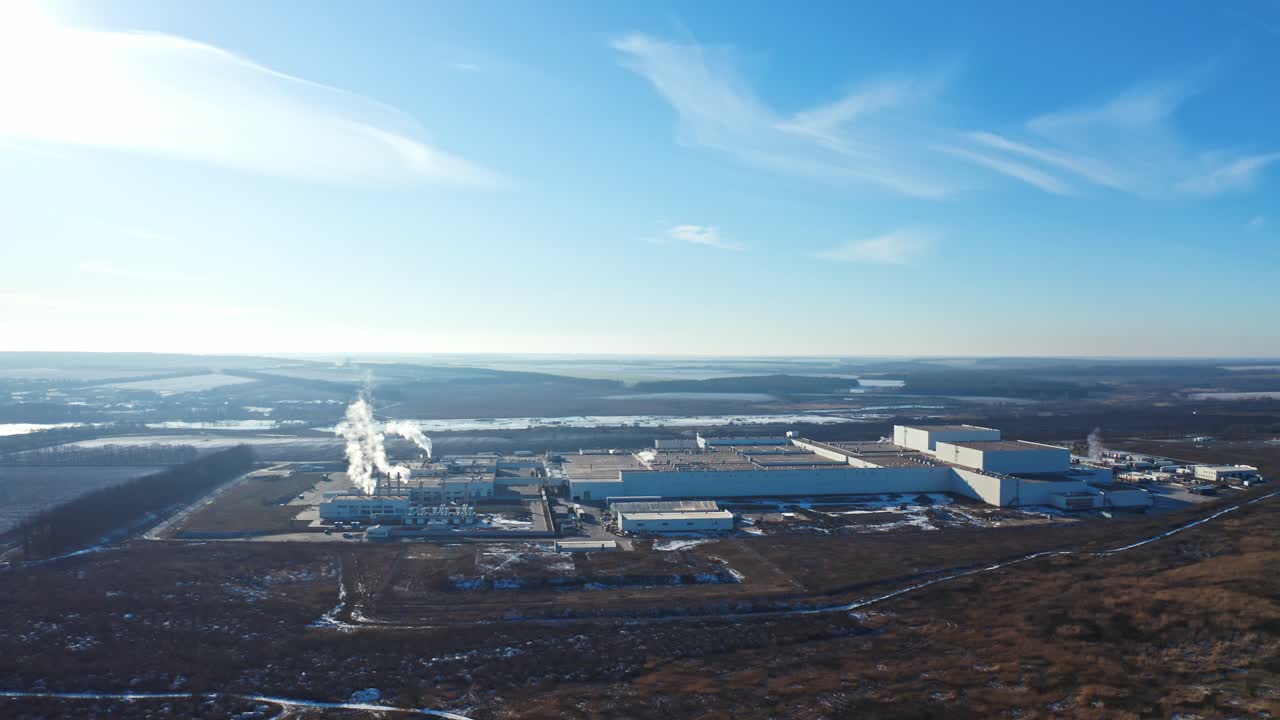 Industrial factory in environment under blue sky. Manufacturing among nature in winter. White smoke coming out of pipes into the air. Aerial view.