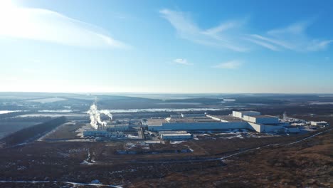 industrial factory in environment under blue sky. manufacturing among nature in winter. white smoke coming out of pipes into the air. aerial view.