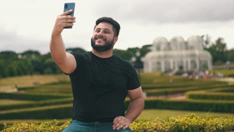 Young-handsome-man-picking-up-his-cell-phone-and-taking-a-selfie-at-a-park-outdoors-while-seated-on-a-bench-located-at-Botanical-Garden,-Curitiba,-Brazil