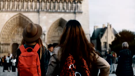 two happy female walking with red backpacks near the notre dame, famous cathedral or church in paris, france