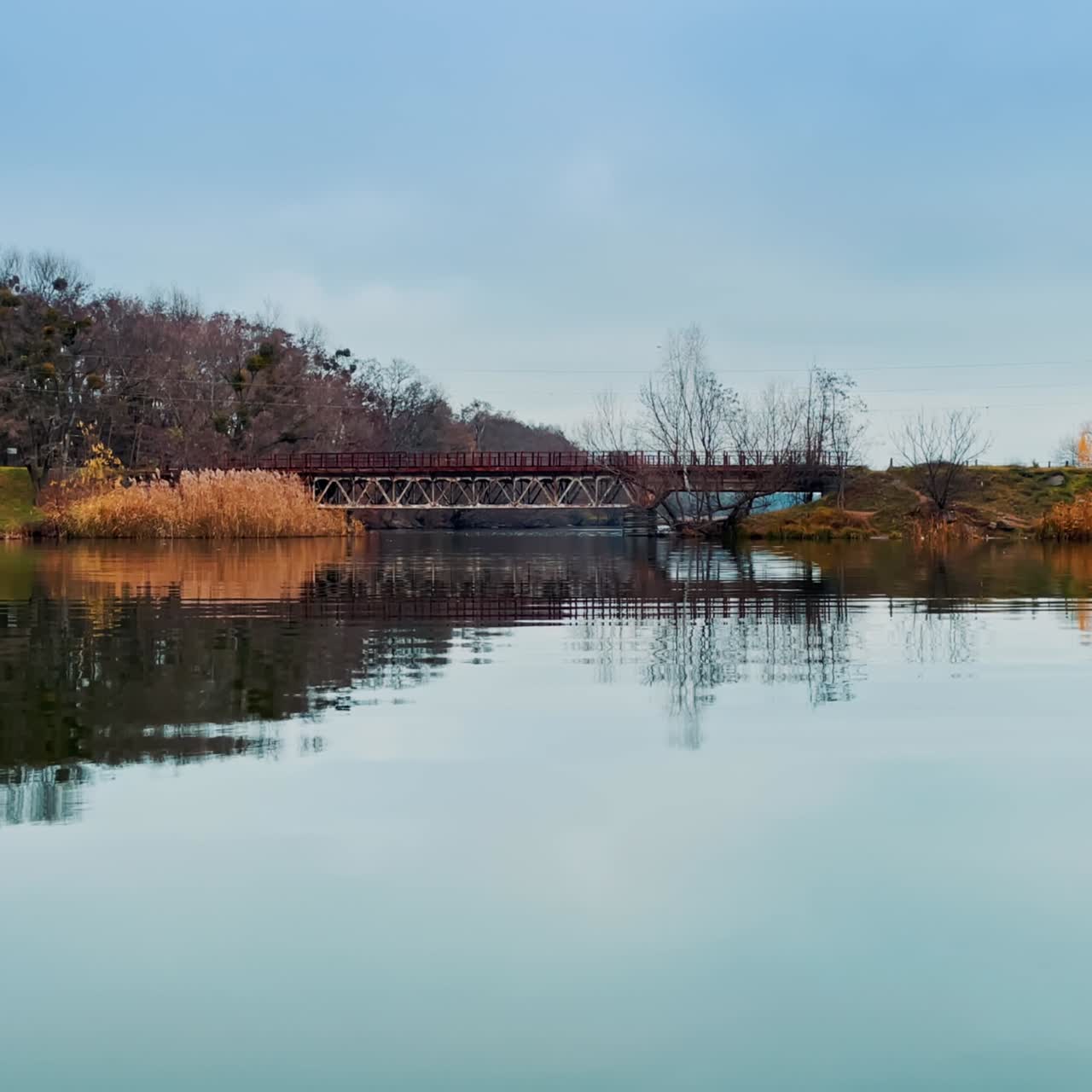 Two dry river banks connected by the metal bridge. Bare trees contaminated by mistletoe plants at backdrop. Blue sky reflecting in the smooth water surface