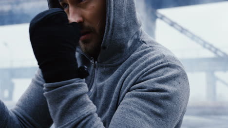 close-up view of caucasian man in grey hoodie hitting a punching bag outdoors an abandoned factory on a cloudy morning