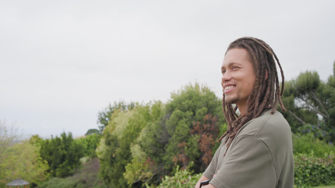 African American man standing scanning in garden, turning, smiling in olive tee, watch, copy space