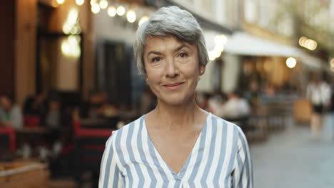 Retrato-De-Una-Hermosa-Anciana-Con-El-Pelo-Gris-Mirando-Directamente-A-La-Cámara-Y-Sonriendo-Alegremente-Al-Aire-Libre-En-La-Ciudad