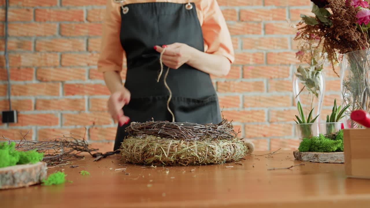Florist in black apron standing at wooden table with brick wall background preparing handmade wreath from dried grass and twigs surrounded by moss,floral arrangements for creative natural decoration project
