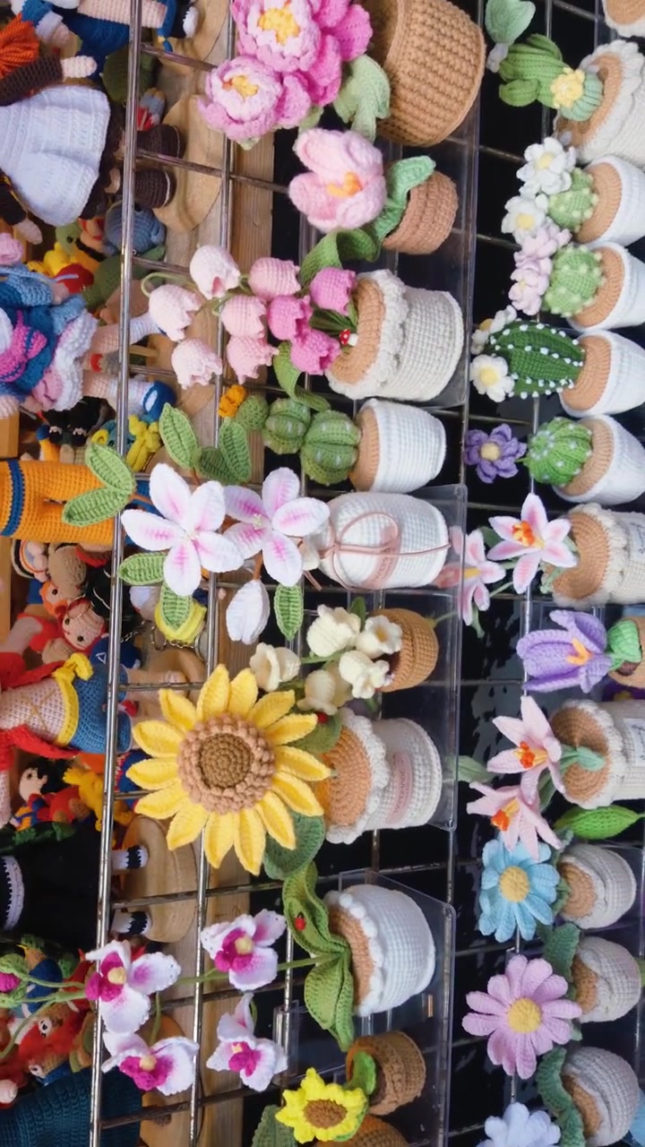 A vertical shot shows colorful crocheted flowers and cacti in small pots arranged on a display rack