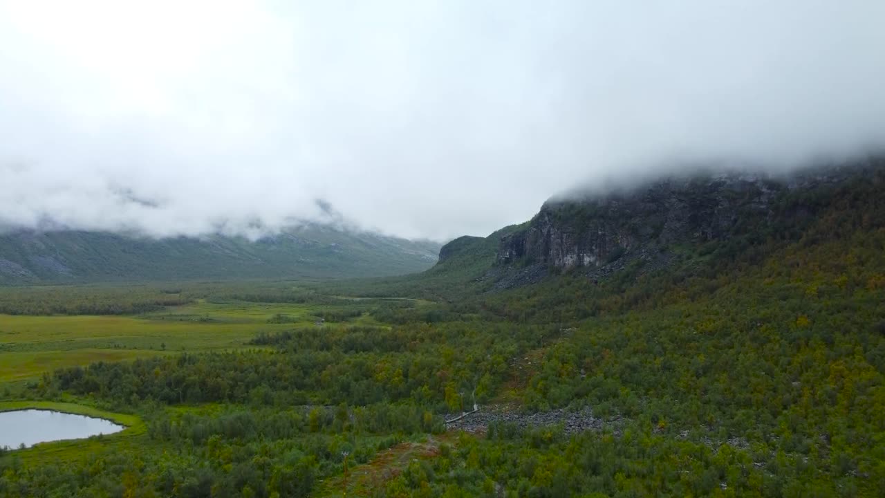 Aerial drone footage rising higher and revealing gorgeous vibrant green and brown Sweden marshland or bog landscapes with reflective cold lakes in front and large mountain in clouds in the background.