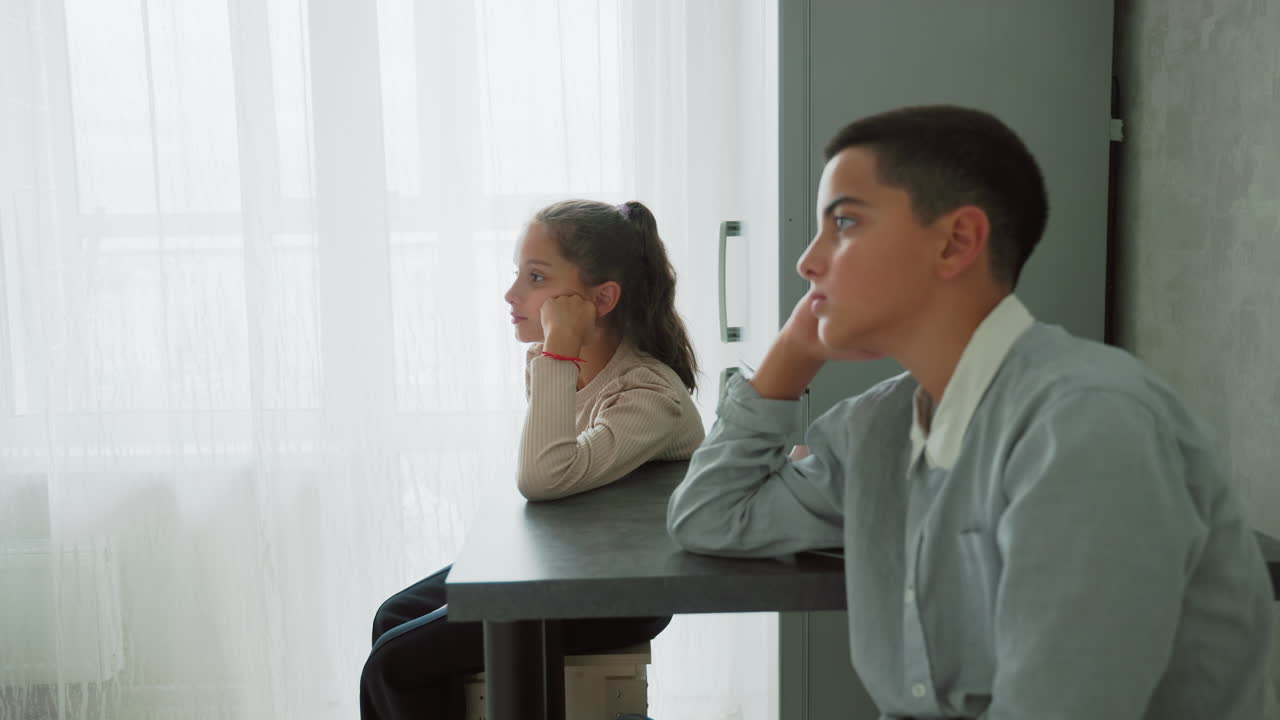 boy and girl in school uniforms sitting quietly at table near bright window waiting patiently in morning light contemplative expressions indoor home setting suggesting early start or preparation scene