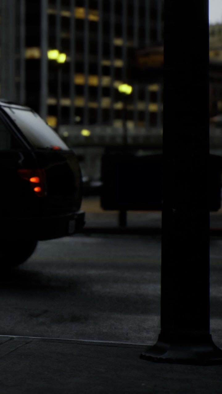 Hombre de pie cerca de un coche negro en una calle urbana de noche con luces alrededor.