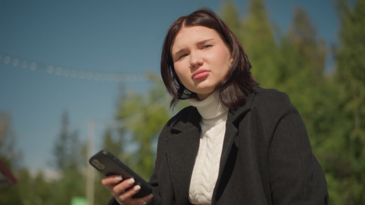 흑색 코트와 색 스웨터를 입고, 밖에서 앉아있는 사려 깊은 여성, 사려깊은 표정으로 멀리보고, 반지 장식 손이 보이고, 배경에는 흐릿한 초록색이 있습니다.