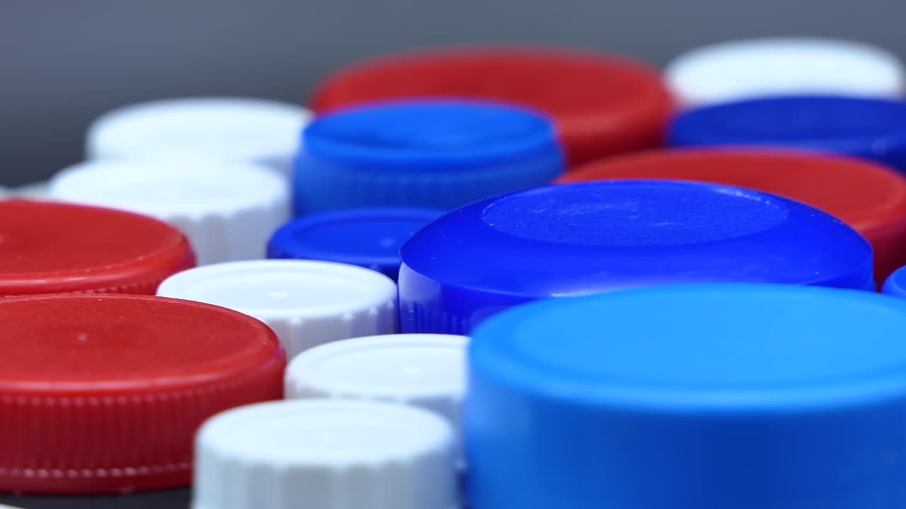 Tappi di plastica usati, colorati di rosso, blu e bianco, che ruotano velocemente davanti a uno sfondo grigio.