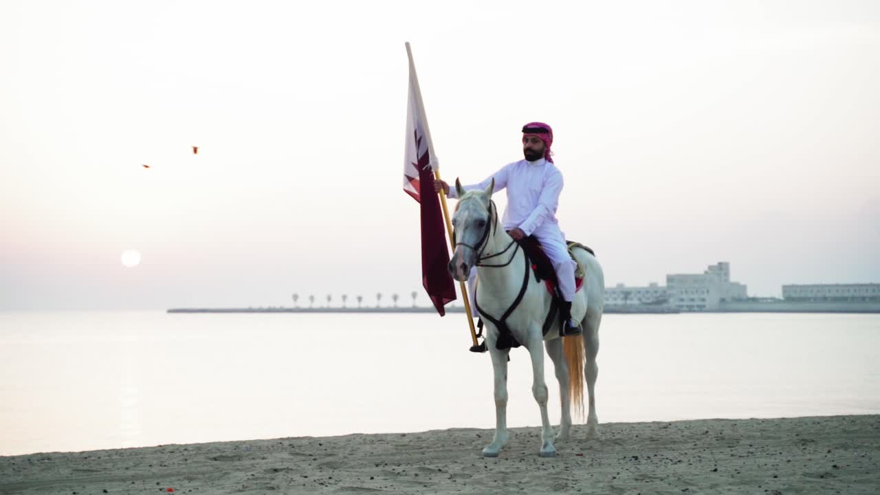 un caballero montando un caballo sosteniendo la bandera de qatar cerca del mar-1
