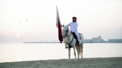 Un-Caballero-Montando-Un-Caballo-Sosteniendo-La-Bandera-De-Qatar-Cerca-Del-Mar-1