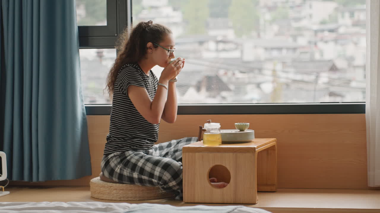 ホテルの部屋で窓辺での朝のティータイム。ゲストがカップを啜る様子、低い木製テーブル、大きな窓から見える街の景色、柔らかなカーテン、居心地の良いクッション席、暖かい日差し。旅への物思いにゆっくりと浸る時間。