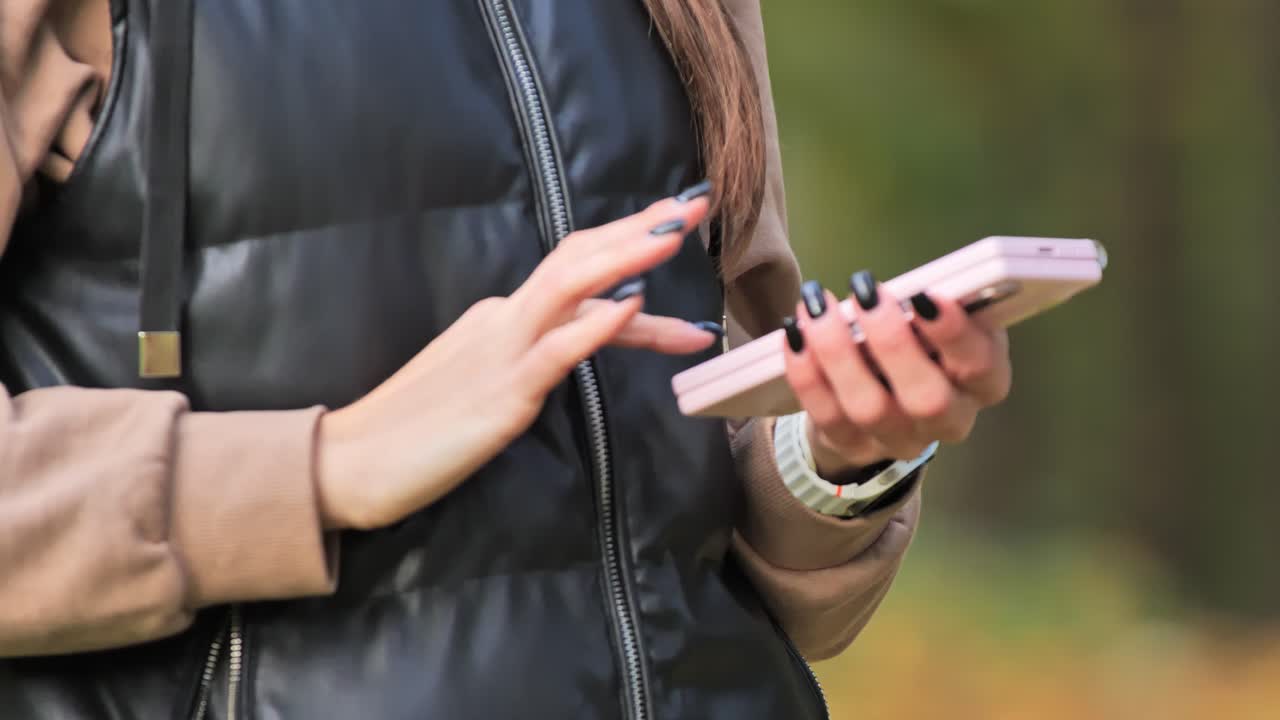 Woman’s hands with dark nails tapping on a smartphone in a green outdoor setting