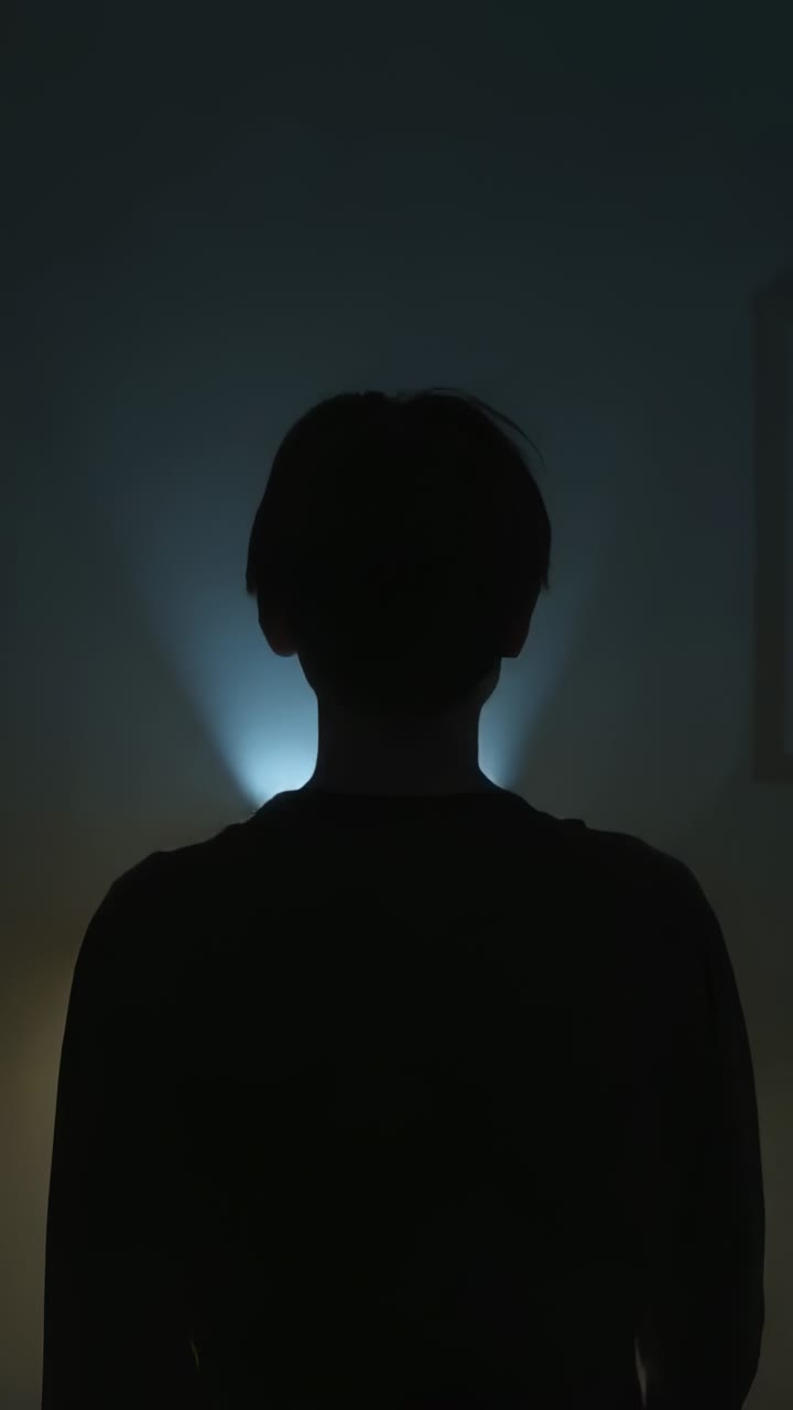 Vertical video: Reacting to blue light brightening, shadowed figure tilting head in dim living room