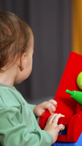 little baby boy in light green shirt holding a big colorful lorry. brown-haired kid playing with a toy in the room and looking around. blurred backdrop. vertical video