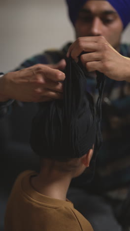 video vertical de primer plano de un padre atando un turbante a la cabeza de un joven sikh, su hijo con el nudo superior sentado en el sofá en casa.
